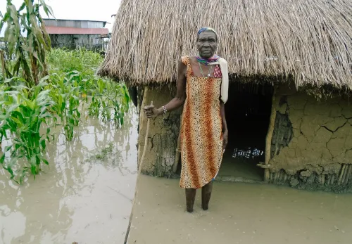 Kriterien: Ältere Dame im Südsudan steht vor ihrem Haus, das bis zum Eingang unter Wasser steht